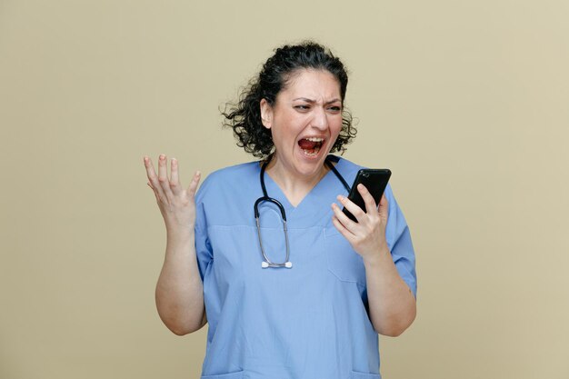 Médica de meia idade com raiva vestindo uniforme e estetoscópio no pescoço segurando o celular olhando para ele mantendo a mão no ar gritando bem alto isolado no fundo verde oliva