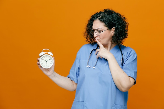 Médica de meia-idade confusa usando óculos uniformes e estetoscópio em volta do pescoço mostrando despertador olhando para ele enquanto toca o lábio isolado em fundo laranja