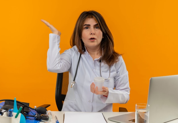 Médica de meia-idade confusa, vestindo bata médica e estetoscópio, sentada à mesa com a área de transferência de ferramentas médicas e laptop segurando um copo médico, mostrando a mão vazia