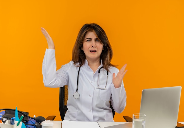 Médica de meia-idade confusa, vestindo túnica médica com estetoscópio, sentada na mesa de trabalho no laptop com ferramentas médicas, levantando as mãos na parede laranja isolada com espaço de cópia