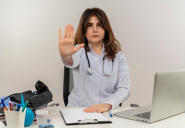 Médica de meia-idade estrita vestindo túnica médica com estetoscópio sentada na mesa de trabalho no laptop com ferramentas médicas mostrando gesto de parada em backgroung branco isolado com espaço de cópia