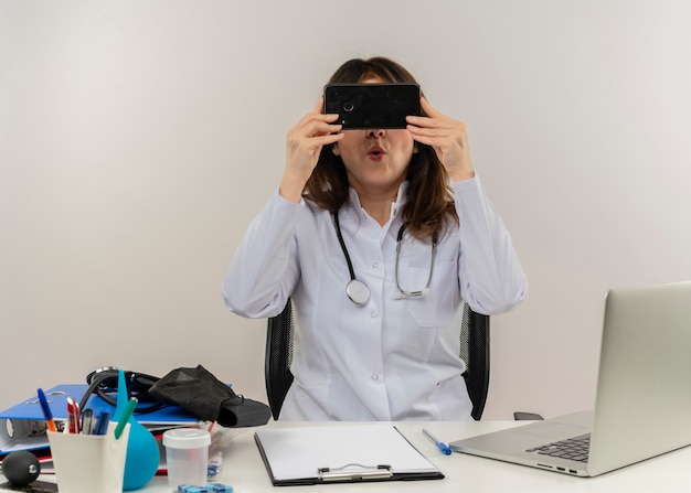 Médica de meia-idade impressionada usando túnica médica e estetoscópio sentada à mesa com a área de transferência de ferramentas médicas e laptop segurando e se escondendo atrás de um telefone celular isolado