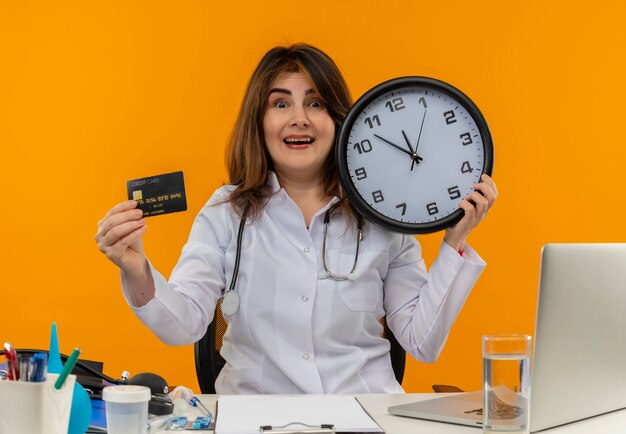 Foto grátis médica de meia-idade impressionada usando túnica médica e estetoscópio sentada na mesa com a área de transferência de ferramentas médicas e laptop segurando o relógio e o cartão de crédito isolados