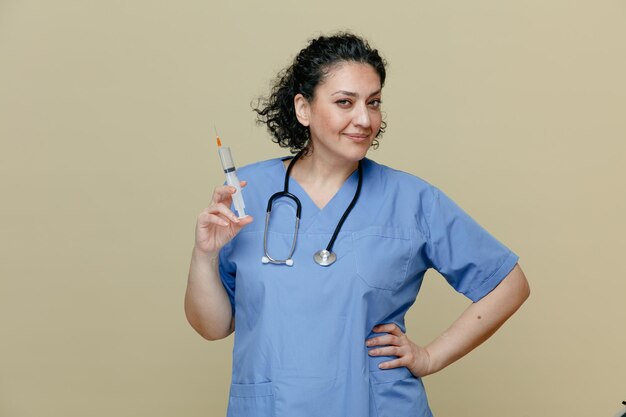 Médica de meia-idade satisfeita e confiante vestindo uniforme e estetoscópio no pescoço segurando a seringa com agulha olhando para a câmera enquanto mantém a mão na cintura isolada no fundo verde-oliva