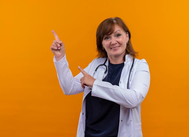 Médica de meia-idade sorridente usando roupão médico e estetoscópio apontando com os dedos para cima na parede laranja isolada com espaço de cópia