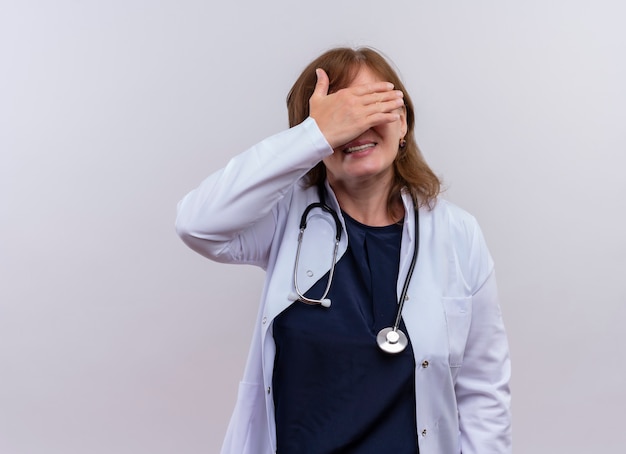 Médica de meia-idade sorridente usando roupão médico e estetoscópio fechando os olhos com a mão na parede branca isolada com espaço de cópia