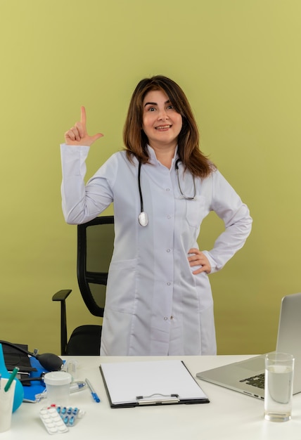 Médica de meia-idade sorridente vestindo túnica médica com estetoscópio em pé atrás de uma mesa de trabalho no laptop com pontos de ferramentas médicas para cima e colocando a mão no quadril na parede verde