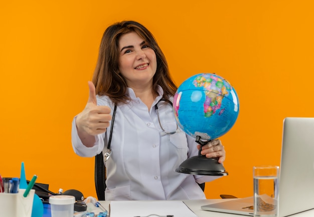 Médica de meia-idade sorridente vestindo túnica médica com estetoscópio sentada na mesa de trabalho no laptop com ferramentas médicas segurando o globo e o polegar para cima em um fundo laranja isolado com espaço de cópia