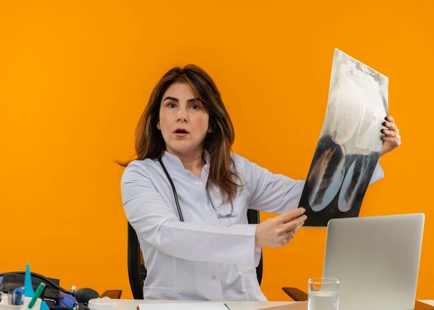 Médica de meia-idade surpresa, vestindo túnica médica com estetoscópio, sentada na mesa de trabalho no laptop com ferramentas médicas, segurando um raio-x em fundo laranja isolado com espaço de cópia