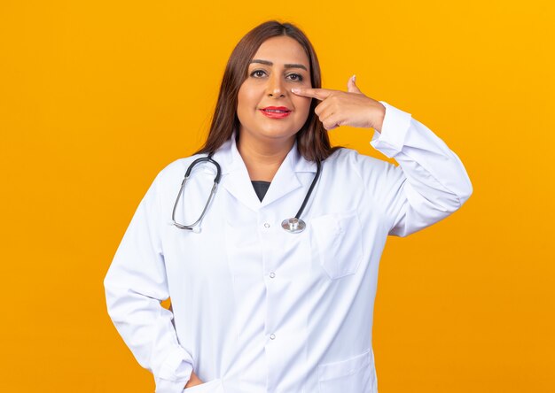 Médica de meia-idade usando jaleco branco com estetoscópio sorrindo apontando com o dedo indicador para o nariz, parada sobre a parede laranja