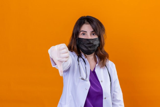 Médica de meia-idade usando jaleco branco com máscara facial protetora preta e estetoscópio olhando para a câmera com rosto sério mostrando o polegar para baixo em pé sobre fundo laranja