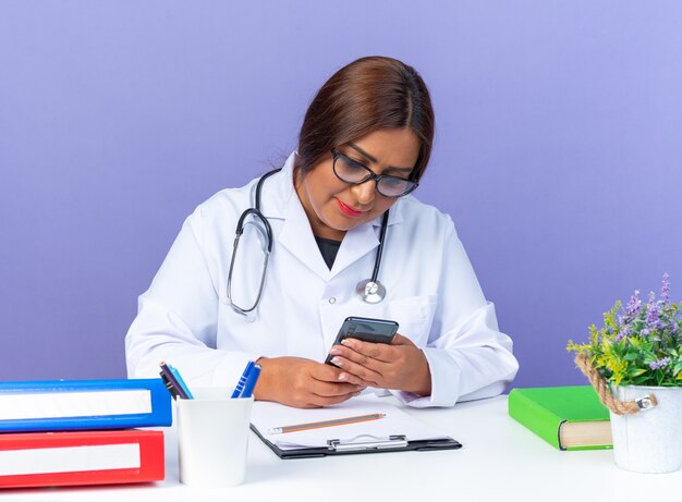 Médica de meia-idade vestindo jaleco branco com estetoscópio usando óculos segurando um smartphone olhando para ele com um sorriso no rosto, sentada à mesa sobre a parede azul