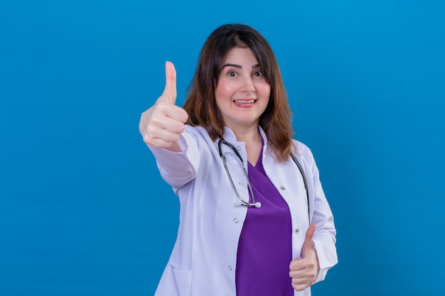 Médica de meia-idade vestindo jaleco branco e com estetoscópio olhando para a câmera, sorrindo alegremente, mostrando os polegares em pé sobre um fundo azul