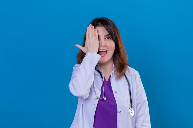 Médica de meia-idade vestindo jaleco branco e com estetoscópio sorrindo e rindo com a mão no rosto cobrindo os olhos para a surpresa em pé sobre um fundo azul isolado