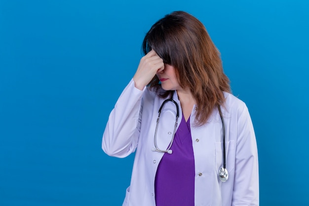 Médica de meia-idade vestindo jaleco branco e com estetoscópio tocando o nariz entre os olhos fechados estressada, sentindo cansaço em pé sobre um fundo azul isolado