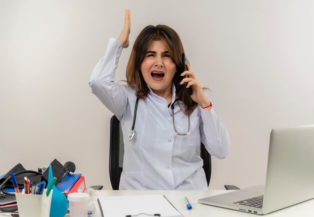 Médica de meia-idade zangada usando um manto médico com estetoscópio sentado na mesa de trabalho no laptop com ferramentas médicas fala no telefone levantando a mão na parede branca com espaço de cópia