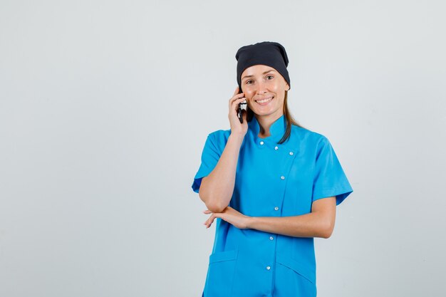 Médica de uniforme azul, chapéu preto, falando no smartphone e parecendo animada