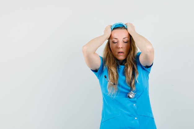 Médica de uniforme azul com as mãos na cabeça e parecendo melancólica