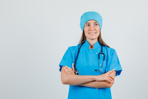 Médica de uniforme azul olhando para longe com os braços cruzados e parecendo esperançosa