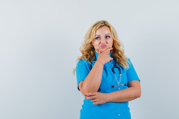 Médica de uniforme azul segurando a mão no queixo e olhando para o espaço pensativo para o texto