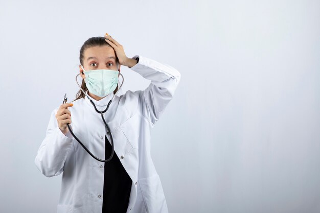 Médica de uniforme branco usando uma máscara médica e segurando um estetoscópio