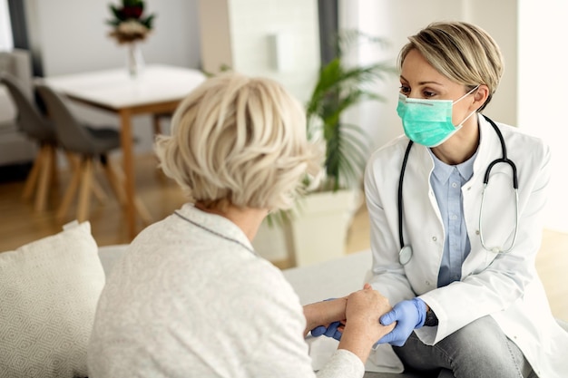 Médica e mulher sênior usando máscaras faciais enquanto seguram as mãos e se comunicam durante a visita domiciliar