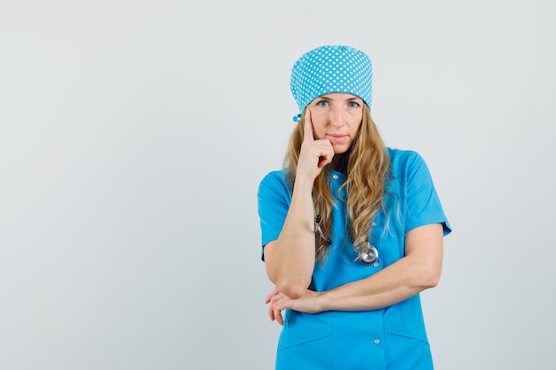 Médica em pé em pose pensativa, de uniforme azul e parecendo inteligente