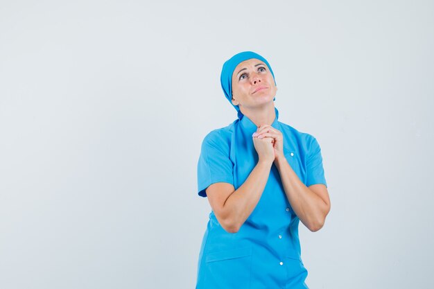 Médica em uniforme azul, apertando as mãos em gesto de oração e olhando esperançosa, vista frontal.