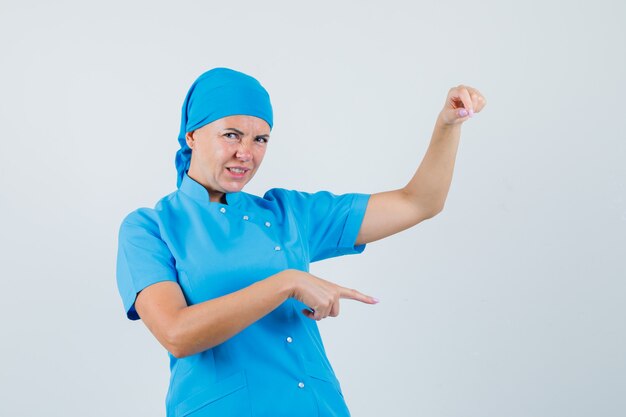 Médica em uniforme azul, fingindo segurar algo, apontando para baixo e parecendo descontente, vista frontal.