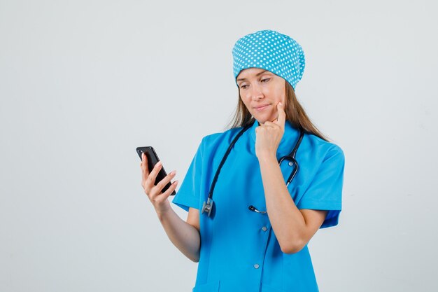 Médica em uniforme azul, usando smartphone com o dedo na bochecha e parecendo um sonho, vista frontal.