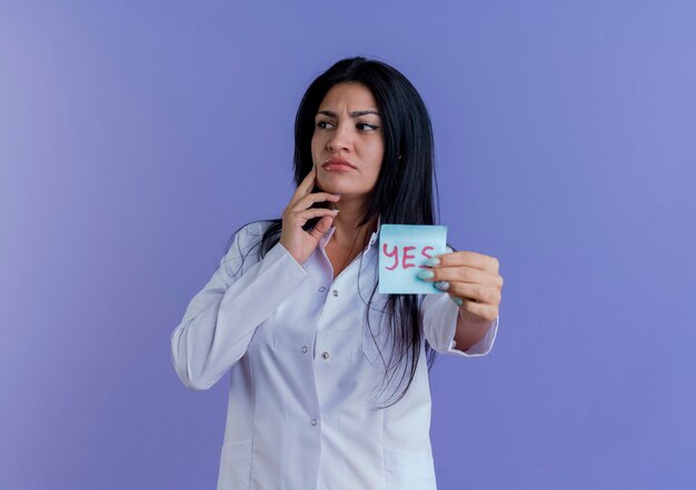 Médica jovem atenciosa vestindo túnica médica mostrando nota sim, olhando para o lado tocando o rosto isolado na parede roxa com espaço de cópia