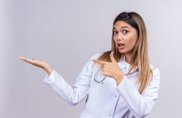 Médica jovem bonita vestindo jaleco branco com estetoscópio, apresentando-se com o braço da mão e apontando com o dedo para o lado, parecendo surpresa