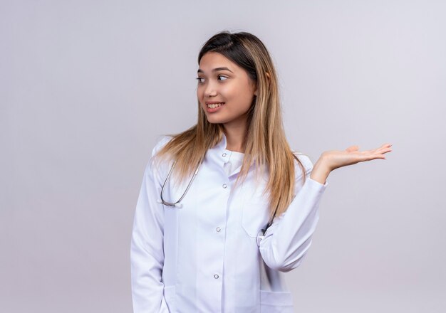 Médica jovem bonita vestindo jaleco branco com estetoscópio apresentando um produto com a mão