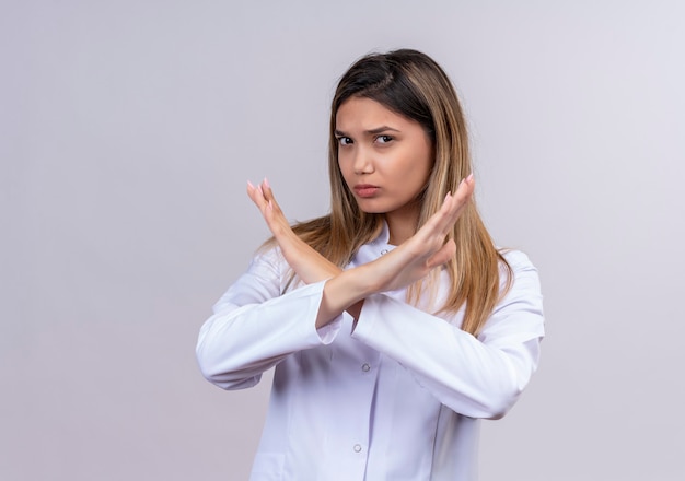 Médica jovem bonita vestindo jaleco branco com estetoscópio e olhando com cara séria fazendo sinal de parada, cruzando os braços