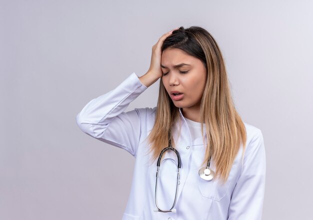 Médica jovem bonita vestindo jaleco branco com estetoscópio e parecendo cansada tocando a cabeça com dor de cabeça