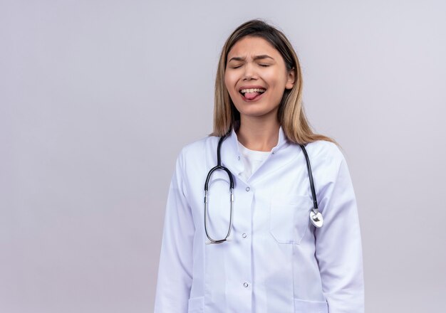 Médica jovem bonita vestindo jaleco branco com estetoscópio em pé com os olhos fechados mostrando a língua com expressão de nojo