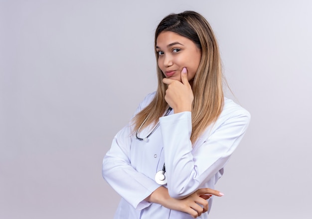 Médica jovem bonita vestindo jaleco branco com estetoscópio olhando com a mão no queixo e expressão pensativa no rosto