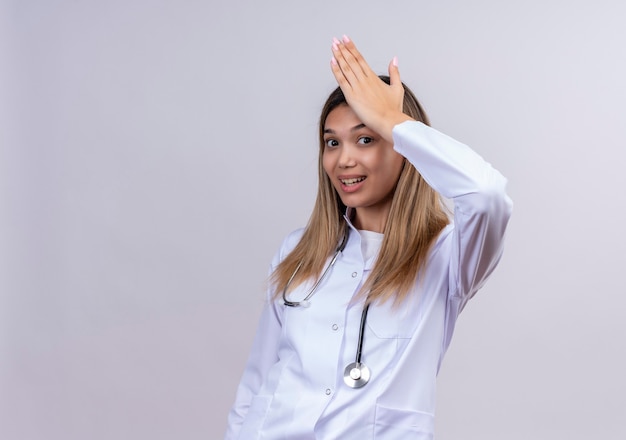 Médica jovem bonita vestindo jaleco branco com estetoscópio, parecendo confusa e incerta, com a mão na cabeça por engano