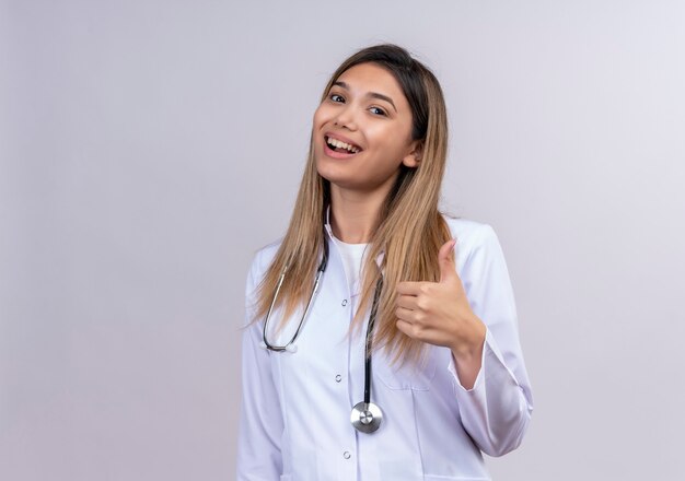 Médica jovem bonita vestindo jaleco branco com estetoscópio, parecendo feliz e animada mostrando os polegares para cima