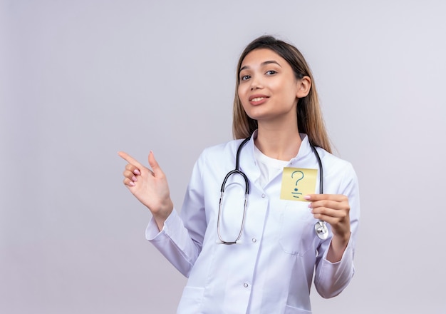 Médica jovem bonita vestindo jaleco branco com estetoscópio segurando um papel lembrete com ponto de interrogação sorrindo alegremente apontando com o dedo para o lado