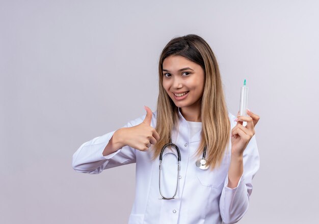 Médica jovem bonita vestindo jaleco branco com estetoscópio segurando uma seringa e sorrindo alegremente mostrando os polegares para cima