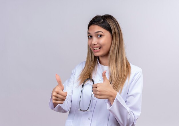 Médica jovem bonita vestindo jaleco branco com estetoscópio sorrindo alegremente mostrando os polegares para cima