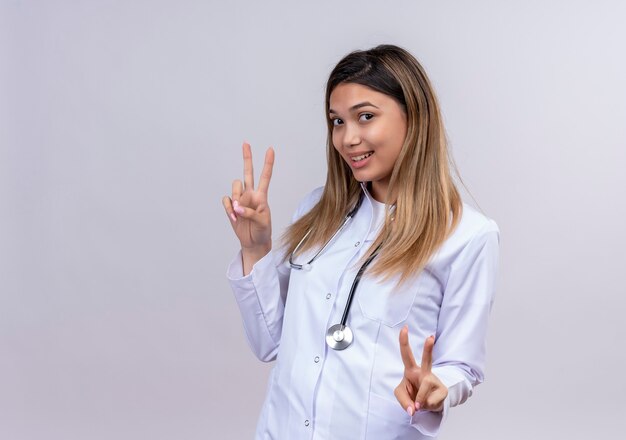 Médica jovem bonita vestindo jaleco branco com estetoscópio sorrindo alegremente mostrando sinais de vitória com ambas as mãos
