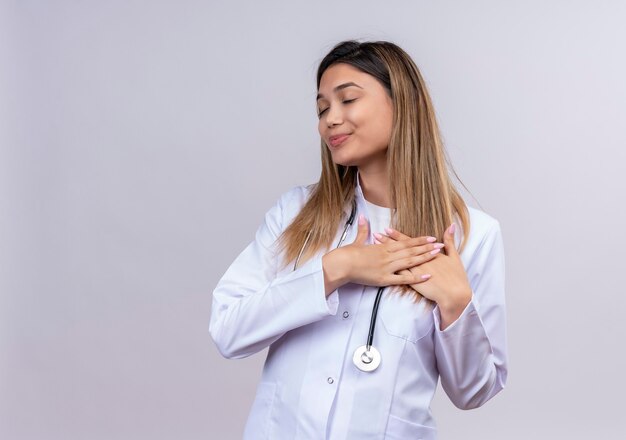 Médica jovem bonita vestindo jaleco branco com estetoscópio sorrindo com as mãos no peito com expressão de gratidão