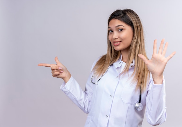 Médica jovem bonita vestindo jaleco branco com estetoscópio sorrindo confiante mostrando o número cinco e apontando com o dedo indicador para o lado