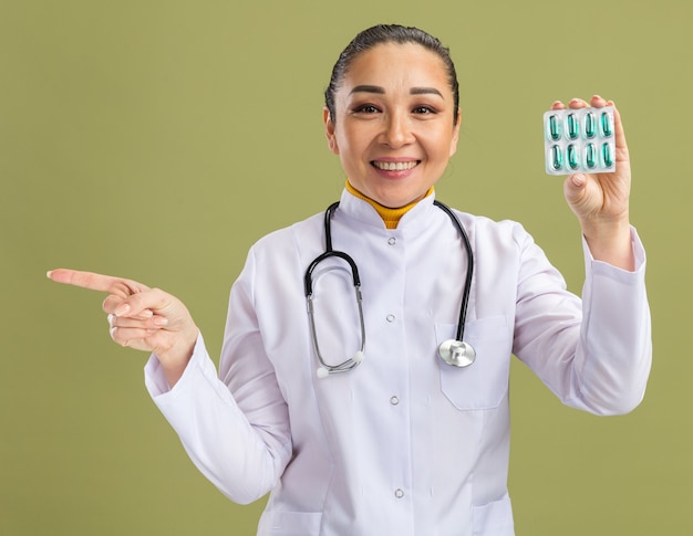 Médica jovem com jaleco branco com estetoscópio no pescoço segurando uma bolha com comprimidos, olhando para a câmera sorrindo confiante apontando com o dedo indicador para o lado na parede verde