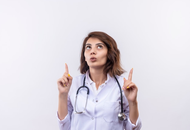 Médica jovem com jaleco branco com estetoscópio olhando para cima surpresa e apontando com dedos finos em pé sobre uma parede branca