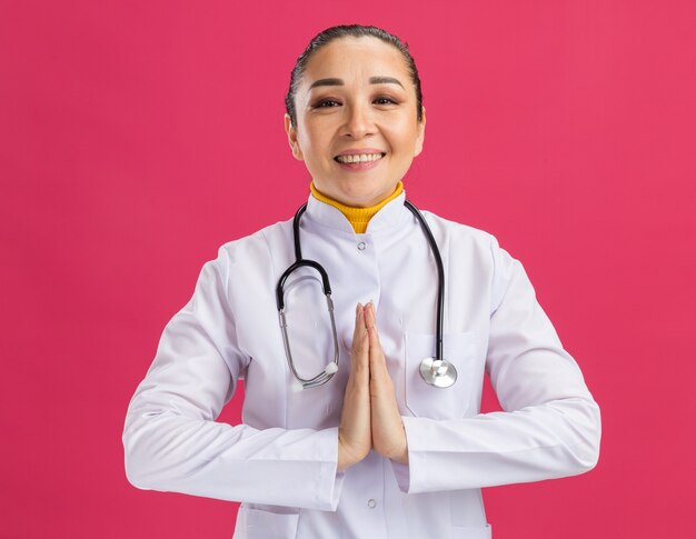 Médica jovem de mãos dadas como um gesto de namastê sorrindo com uma cara feliz