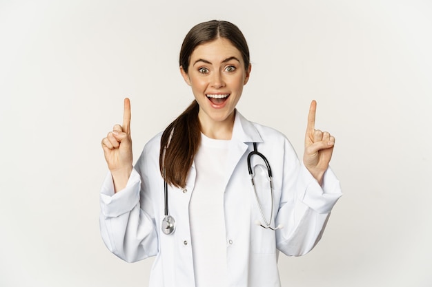 Médica jovem entusiasmada sorrindo, apontando os dedos para cima, vestindo uniforme hospitalar, de pé sobre fundo branco