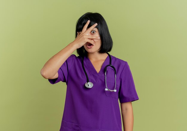 Médica jovem morena assustada de uniforme com estetoscópio coloca a mão no rosto e olha através dos dedos isolados em um fundo verde oliva com espaço de cópia